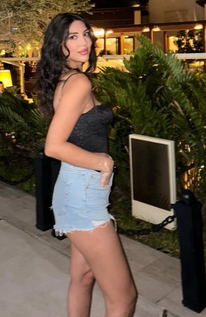 Woman with dark hair wearing a black spaghetti-strap top and ripped blue denim shorts, posing outdoors at night near greenery.